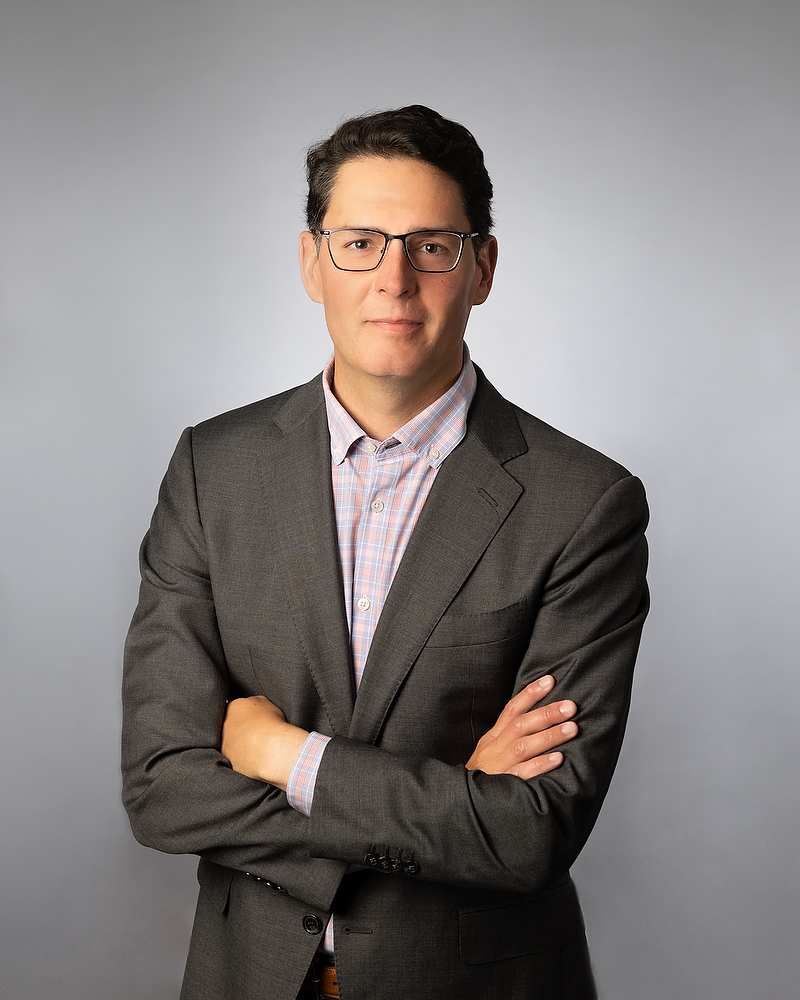 Full body professional headshot of tan white man wearing glasses in a grey suit with are crossed in front of a grey background that is lit taken in a photography studio