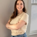 headshot of white woman with brown hair wearing beige top and jeans standing against a wall with arms crossed
