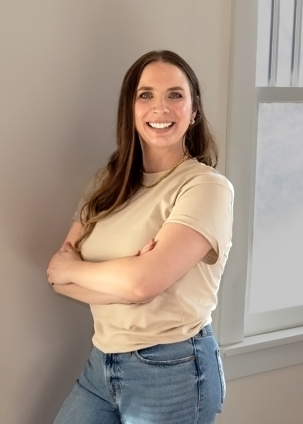 headshot of white woman with brown hair wearing beige top and jeans standing against a wall with arms crossed