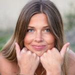 senior portrait of girl with brown hair and blue eyes smirking with fists under chin and thumbs out in each direction